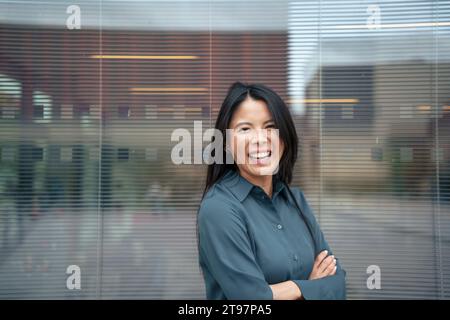 Happy businesswoman standing with arms crossed in front of glass Stock Photo