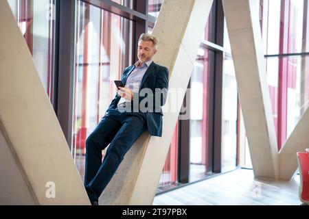 Mature businessman using smart phone and leaning on column Stock Photo