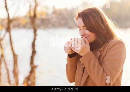 Smiling woman drinking tea from cup by lake at sunset Stock Photo