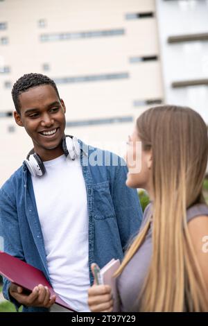 Multiracial couple smiling and holding folders in a campus.Two young students looking each other laughing and relaxed in a park. Stock Photo