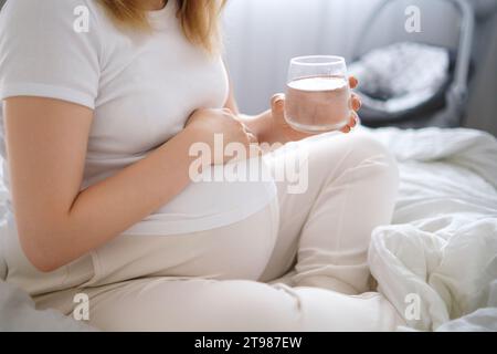 Pregnant woman sits on the bed, tenderly embracing her growing belly and holding glass of water. Stock Photo