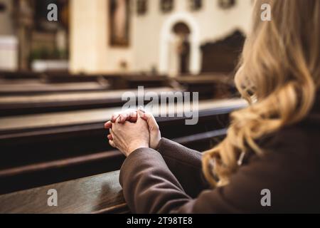 Christian woman with hands folded in prayer sitting on church pew. Religion and spirituality concept. Female person is praying to God Stock Photo