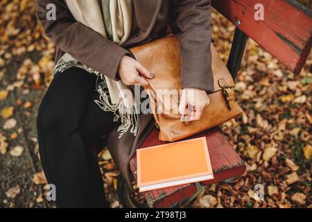 Woman opening leather handbag on bench in autumn park. Relax with book outdoors. Fashion personal accessory Stock Photo