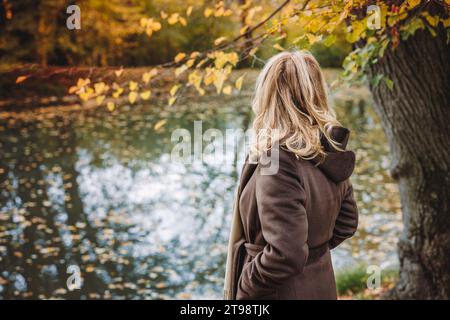 Pensive woman standing at lake in autumn park. Lonely sad female person looking at reflection on water surface Stock Photo