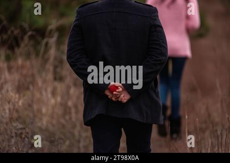 Young man holds red wedding box with ring behind his back. Authentic engagement outdoors in the forest. Winter wedding. Autumn surprise. Lifestyle. Fa Stock Photo