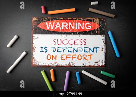 Success is Dependent on Effort. Metal warning sign and colored pieces of chalk on a dark chalkboard background. Stock Photo