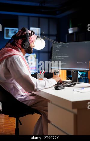 Data developer from the Middle East types code on a computer, demonstrating the core of software programming and development. Muslim man using wireless headphones and immersed in algorithm execution. Stock Photo