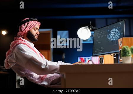 Side-view shot of a young man in Islamic attire analyzing code on a computer screen using AI and machine learning. The image depicts cyber security and data monitoring in a cloud-based system. Stock Photo