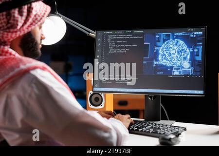 Muslim engineer focuses on a computer screen displaying code and data in a futuristic work setting. He manages a robust cloud-based system while monitoring data on a desktop computer screen. Stock Photo