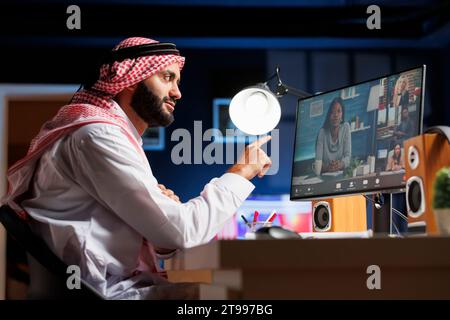 On computer, Muslim guy is carefully listening to his coworkers during group video conversation. Side-view photo shows a Middle Eastern man dressed traditionally holding an online meeting. Stock Photo