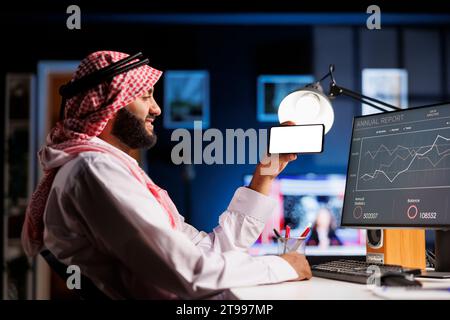 An Arabic individual, wearing traditional clothing uses a mobile phone at an office desk. The isolated white screen offers space for customized content in an advertising or promotional setting. Stock Photo