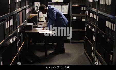 African American policeman carefully analyzes material, does records searches, and collects data from shelves and laptop. Male police officer standing, looking through case files on desk in office. Stock Photo