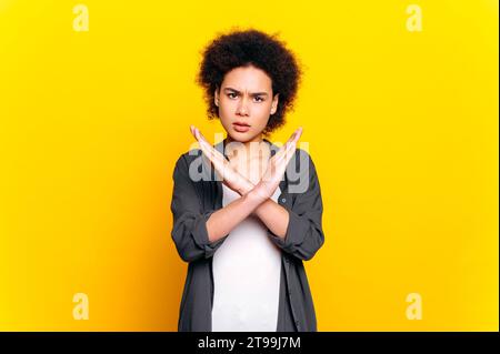 Dissatisfied african american curly haired young woman, shows stop gesture with two hands, against female violence, racism, abuse, stands on isolated yellow background. Stop gesture, disagreement, No Stock Photo