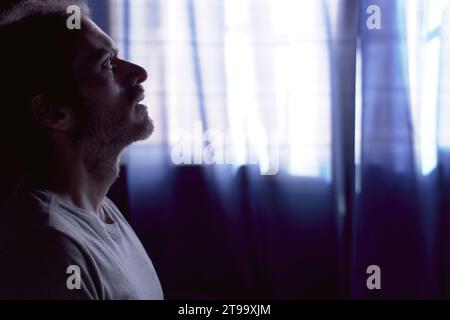 Depressed young bearded man crying in dark room. Dim light with window behind. Concept of depression and mental health, grief and loneliness. Stock Photo