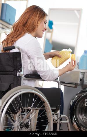 woman in wheelchair by washing machine pouring fabric conditioner Stock Photo