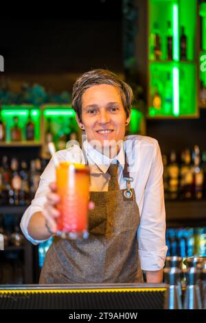 Vertical portrait of a happy bartender serving a luxury cocktail in the counter of a bar Stock Photo