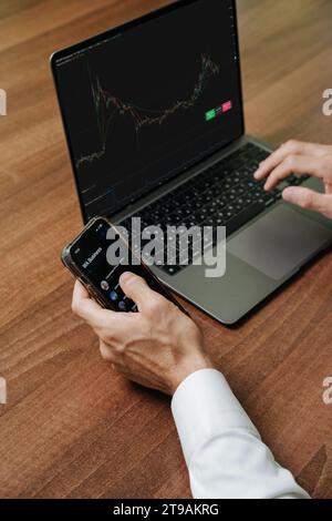 A person sitting at a table where he is engaged in work activities. He has a laptop in front of him, which is displaying a chart suggestive of a stock Stock Photo