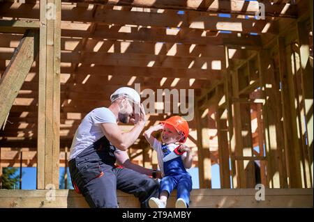 Father with toddler son building wooden frame house. Man giving high five to kid, having fun on the edge of balcony on construction site, wearing helmets and overalls. Carpentry and family concept. Stock Photo