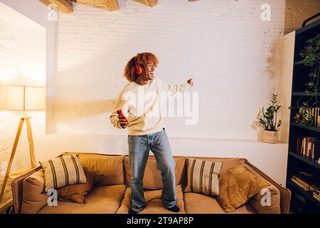Young african american man listening music with red headphones and dancing on a sofa in living room. Very effusive man singing and dancing at home. Stock Photo