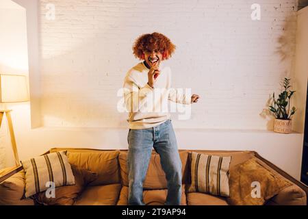 Young african american man listening music with red headphones and dancing on a sofa in living room. Very effusive man singing and dancing at home. Stock Photo