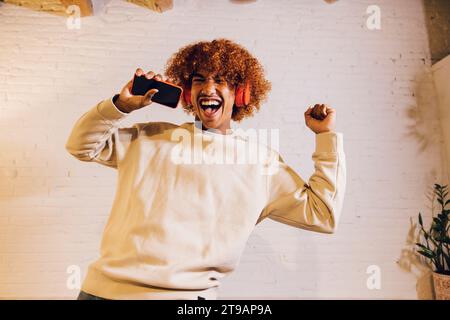Young african american man listening music with red headphones and dancing on a sofa in living room. Very effusive man singing and dancing at home. Stock Photo