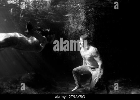 Black and white underwater image of a man and woman swimming in their wedding clothes in a cenote. Stock Photo