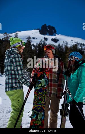 Three friends (one male and two females) hanging out with skis and a snowboard in front of a ski resort. Stock Photo