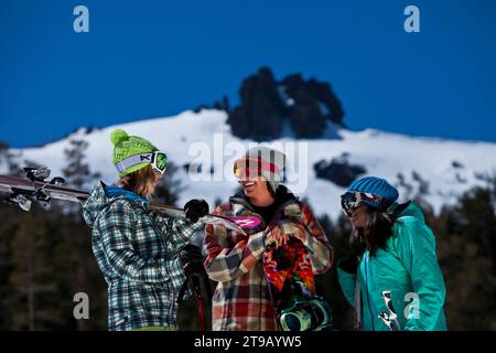 Three friends (one male and two females) hanging out with skis and a snowboard in front of a ski resort. Stock Photo