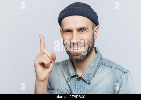 Smiling bearded man in a beanie pointing upwards, wearing a denim shirt against a light background Stock Photo
