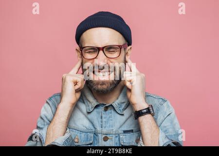 Man in beanie and glasses grimacing with hands on ears, dressed in denim, against a pink backdrop Stock Photo