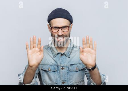 Smiling man in beanie and glasses showing his palms forward in a stopping gesture, against a light grey background Stock Photo