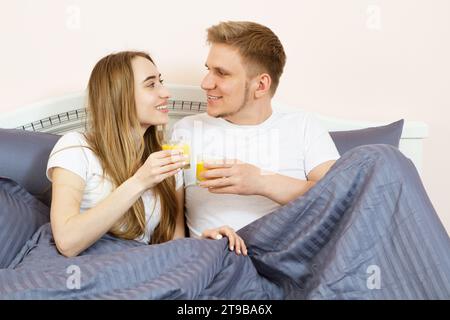 Happy relaxed young couple with orange juice in bed at home. Romantic couple drinking orange juice lying on their bed. Stock Photo