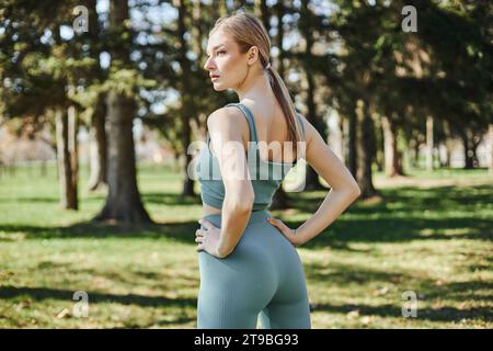 sportive young woman with blonde hair and active wear standing with hands on hips in green park Stock Photo
