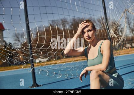 blonde young woman with ponytail in activewear resting while sitting on haunches after workout Stock Photo