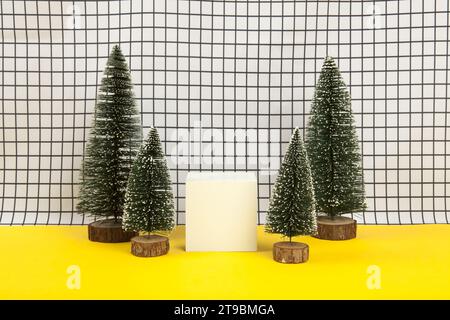 a white wooden cube surrounded by several miniature christmas trees on a black and white grid background. empty object display stand. Minimal still li Stock Photo