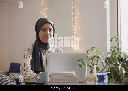 Portrait of young female freelancer wearing hijab and sitting with laptop in living room Stock Photo