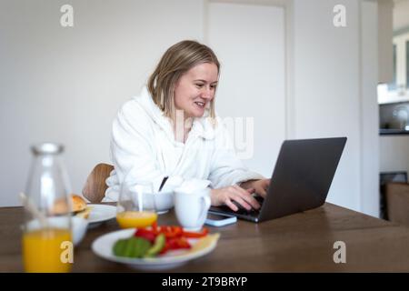 Smiling blond woman using laptop at dining table Stock Photo