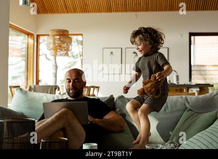 Mature male freelancer working over laptop while his son jumps on sofa and enjoys himself at home Stock Photo