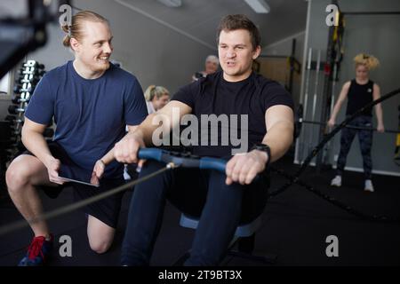 Smiling male instructor encouraging man exercising on rowing machine at gym Stock Photo