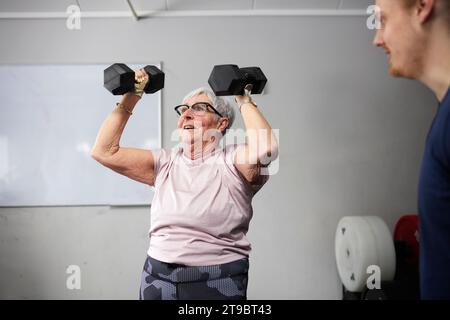 Senior woman exercising with dumbbells by male fitness instructor in gym Stock Photo