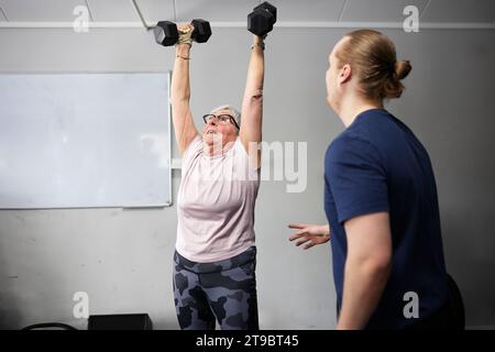 Fitness instructor standing near senior woman exercising with dumbbells in gym Stock Photo