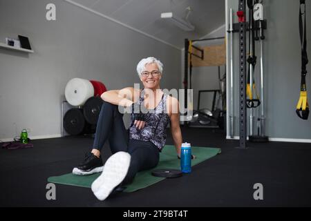 Full length portrait of smiling senior woman sitting on exercise mat at health club Stock Photo