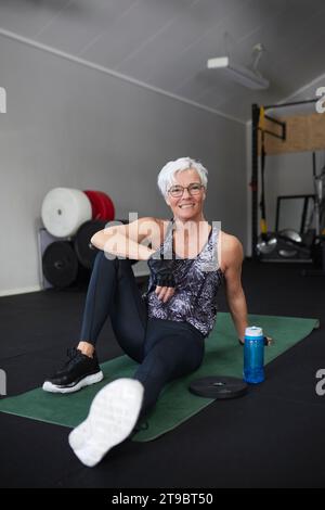 Full length portrait of happy senior woman sitting on exercise mat at health club Stock Photo