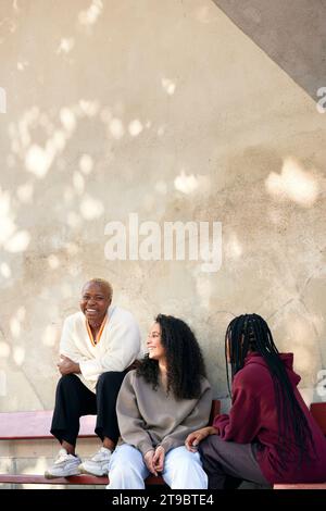 Cheerful young female friends sitting on bench against wall Stock Photo