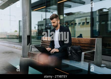 Businessman using laptop sitting on bench while waiting at railroad station Stock Photo