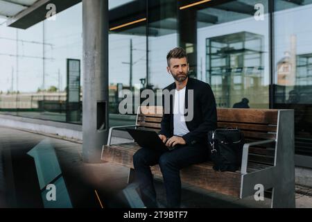 Businessman sitting with laptop on bench while waiting at railroad station Stock Photo