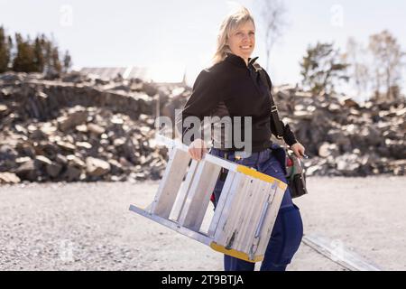 Smiling female electrician carrying ladder while walking on road Stock Photo