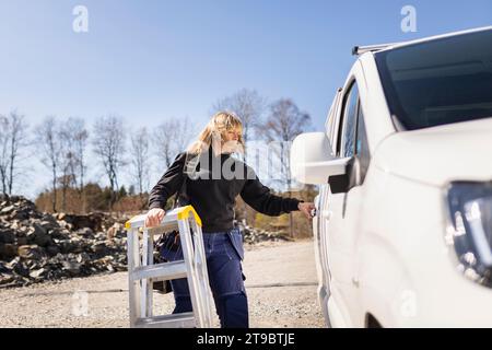 Female electrician with ladder opening van's door standing on sunny day Stock Photo