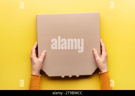 Pizza closed carton box in caucasian hands on uniform yellow background flat lay mockup with blank space Stock Photo