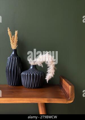 Two black vases with dried grass and plants on a wooden table against a green wall Stock Photo